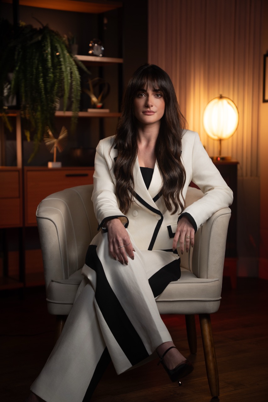 Dra. Ana Paula Bovo sentada na poltrona creme, de terno branco, olhando para a câmera com expressão séria.
