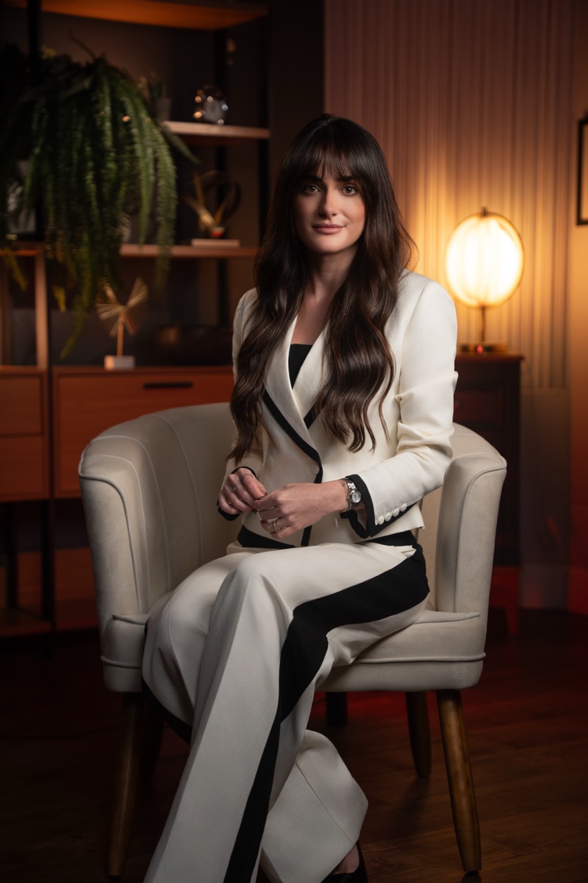 Dra. Ana Paula Bovo sentada na poltrona creme, de terno branco, com um sorriso leve e as mãos no colo.